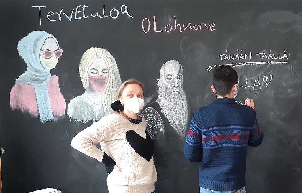 Nainen ja mies liitutaulun luona. Siinä on teksti tervetuloa Olohuone.