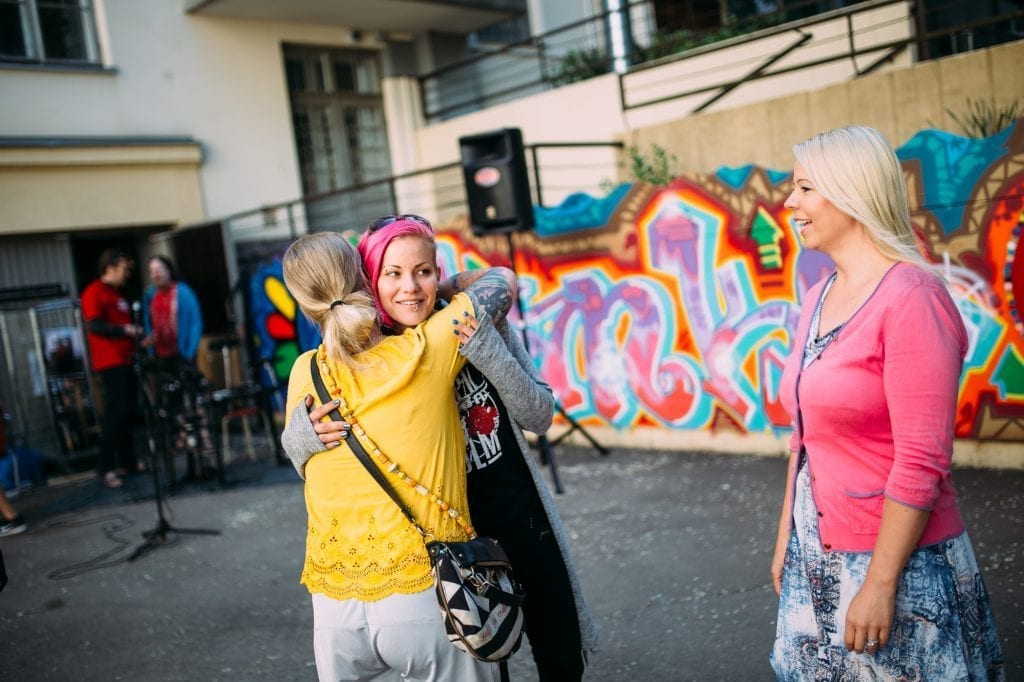 Kaksi naista halaa toisiaan, kolmas seisoo heidän vieressään. Takana on värikäs graffiti.