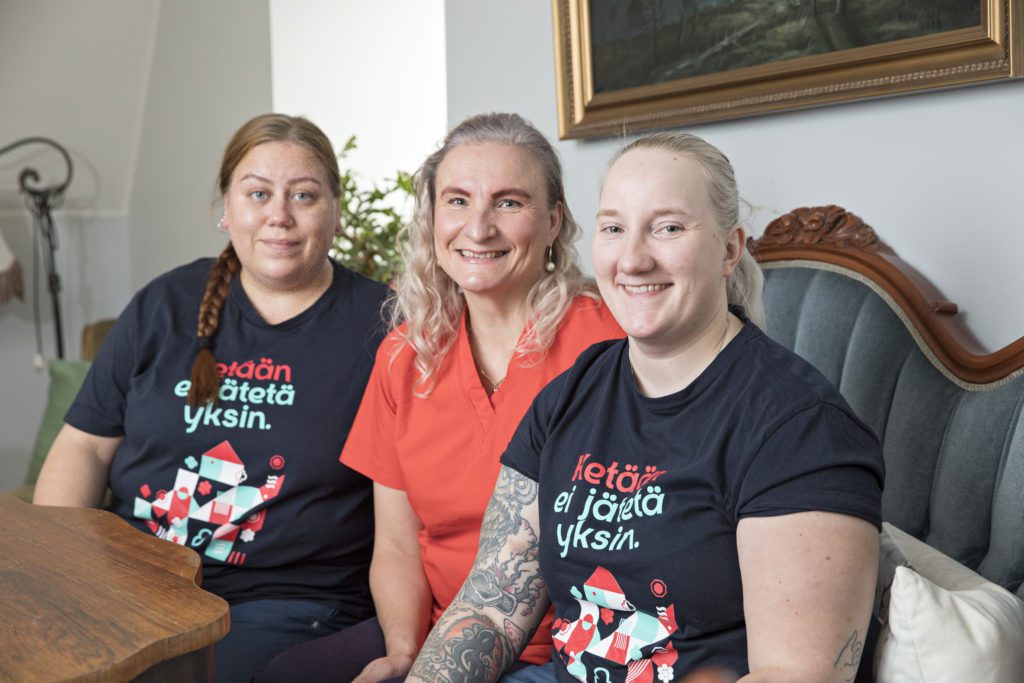Kolme naista istuu vierekkäin ja katsoo kameraan, kahden paidassa teksti "Ketään ei jätetä yksin".
