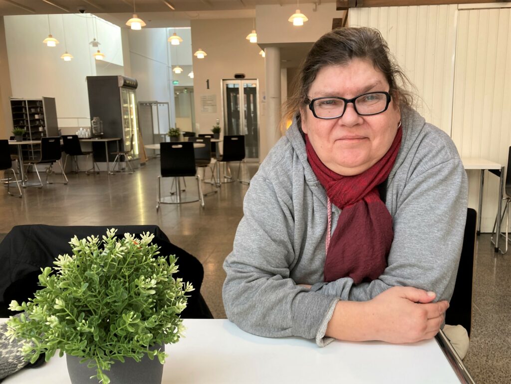 Nainen istuu ravintolan pöydässä ja katsoo kameraan.