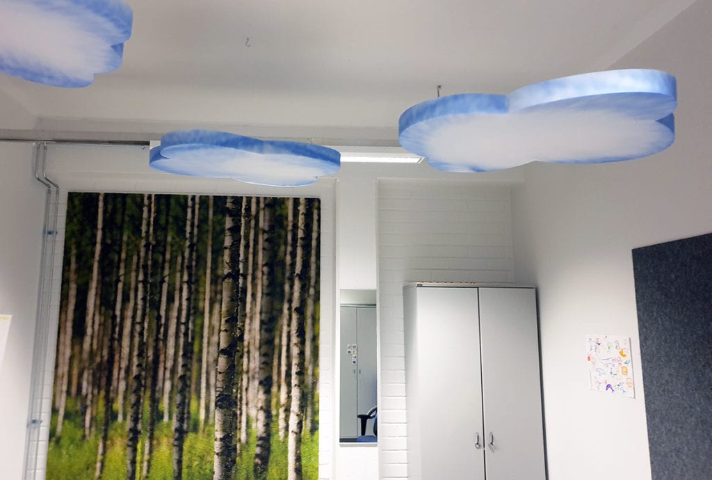 Huoneen katosta roikkuu sinisen ja valoisen sävyisiä, pilvenmuotoisia akustiikkapilviä. Seinällä iso seinän peittävä taulu, jossa kuvana metsämaisema. Huoneessa myös korkeahko seinäkaappi.