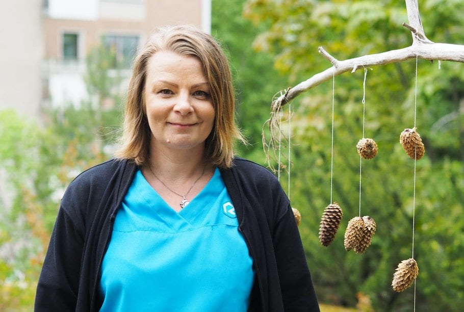 Sinipaitainen ja mustatakkinen nainen seisoo vehreän kuusimetsän edustalla ja katsoo kameraan päin hymyillen.