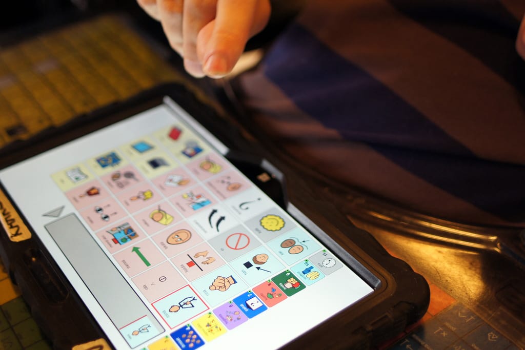 Käsi osoittaa tablettia, jonka ruudulla näkyy kuvakommunikaatiokuvia.
