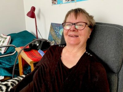 Nainen istuu harmaassa nojatuolissa ja hymyilee.